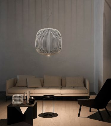Modernes, minimalistisches Wohnzimmer mit einem beigen Sofa, schwarzen Beistelltischen und einer skulpturalen Hängeleuchte aus vertikalen Metallstreben, die Schatten an die Wand wirft.