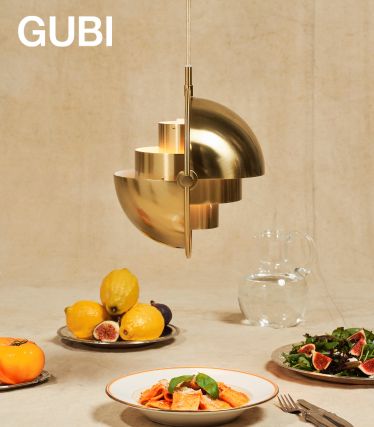 Elegante goldene Pendelleuchte über einem Esstisch mit Tellern voller Speisen, Obst und Wasserkrug, vor neutralem Hintergrund mit dem Logo „GUBI“.