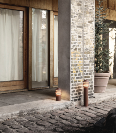 Außenaufnahme mit zwei modernen zylindrischen Wegeleuchten, die neben einem Steinweg und einer Säule aus Ziegeln und Beton aufgestellt sind. Dahinter befinden sich Holzrahmen-Glastüren mit Lichtvorhängen und eine große immergrüne Topfpflanze. Das warme Licht der Leuchten schafft eine gemütliche und einladende Atmosphäre.